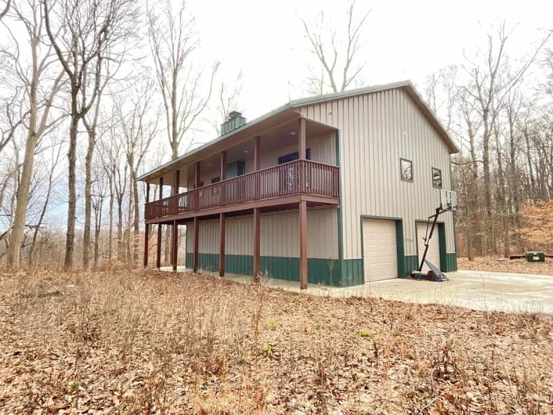 Front exterior of 12144 Fertig Rd in Poland, Indiana: a two-story home with a wraparound deck above a drive-under garage, surrounded by woods.