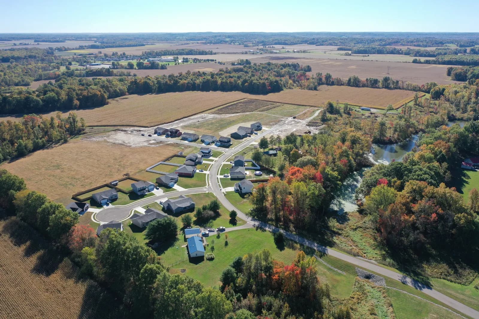 Wide autumn drone aerial over the whole Four Oaks subdivision in Spencer, Indiana, with the creek and farmland beyond.