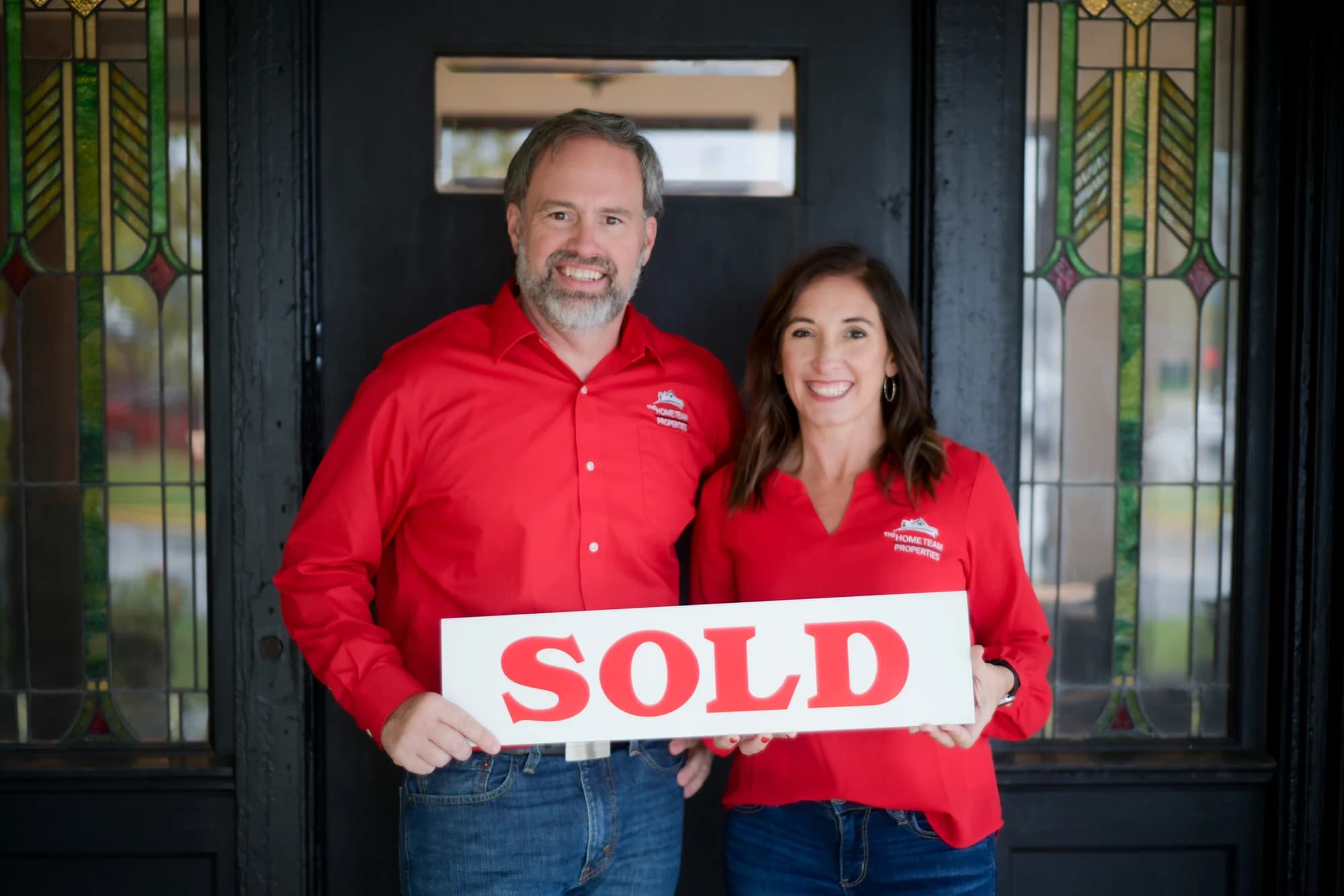 Rob and Samantha of The HOME TEAM Properties holding a red SOLD sign at their Spencer office.