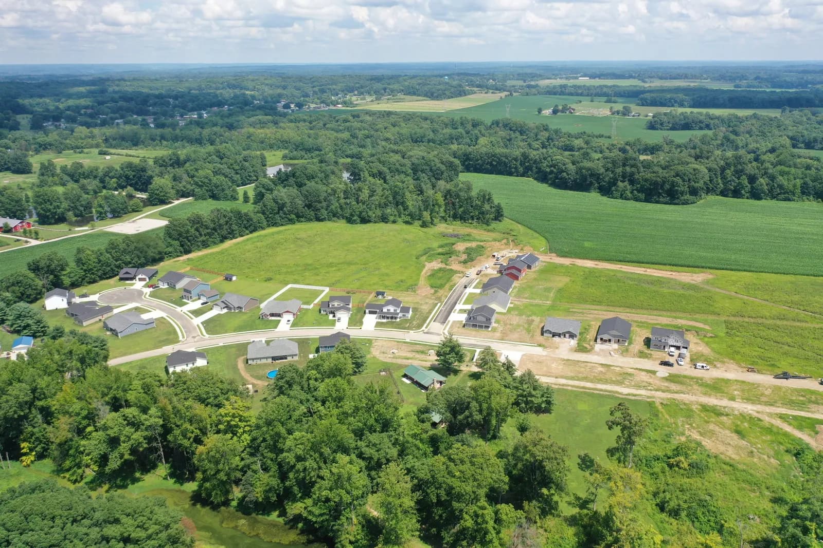 Summer drone aerial over the Four Oaks neighborhood in Spencer, Indiana: finished homes along curving streets, green fields, and the tree line beyond.